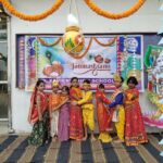 Group of children in colorful traditional indian outfits posing in front of a janmashtami banner at a school celebration with orange marigold garlands overhead and festive decor